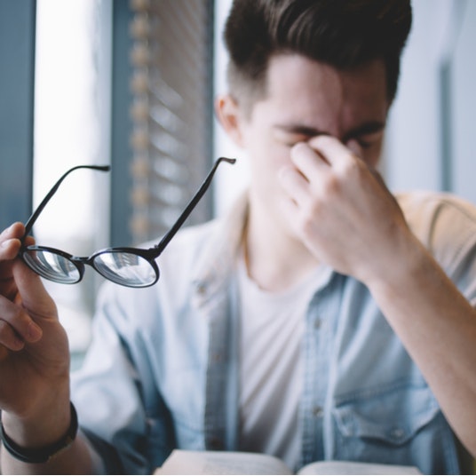 a young man with glasses in one hand and rubbing the bridge of his nose in exhaustion with the other hand