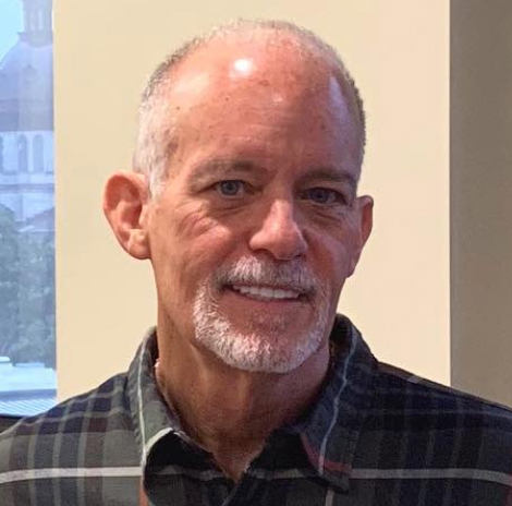 Smiling man with a trimmed white beard wearing a plaid shirt, standing indoors near a window