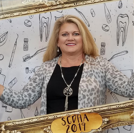Smiling woman with blonde hair holding a decorative photo frame labeled ‘SCDHA 2019,’ standing in front of a dental-themed backdrop