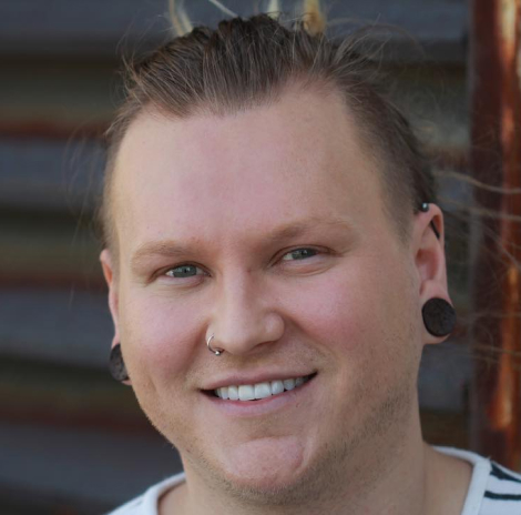 Smiling man with long blond hair tied back, wearing gauges and a nose ring, standing outdoors in front of a metal wall