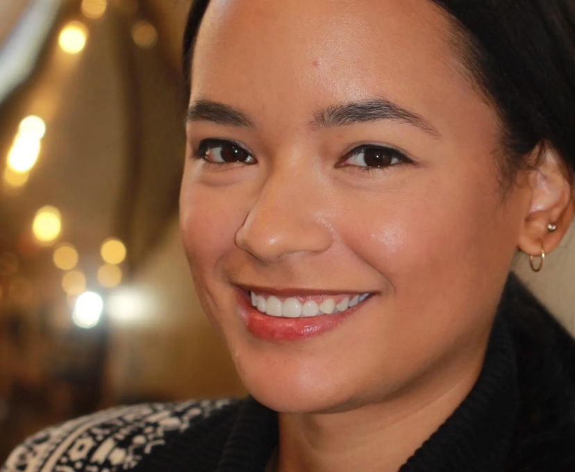 Smiling woman wearing a jeweled tiara, black gloves, and a white sleeveless top posed against a light gray background