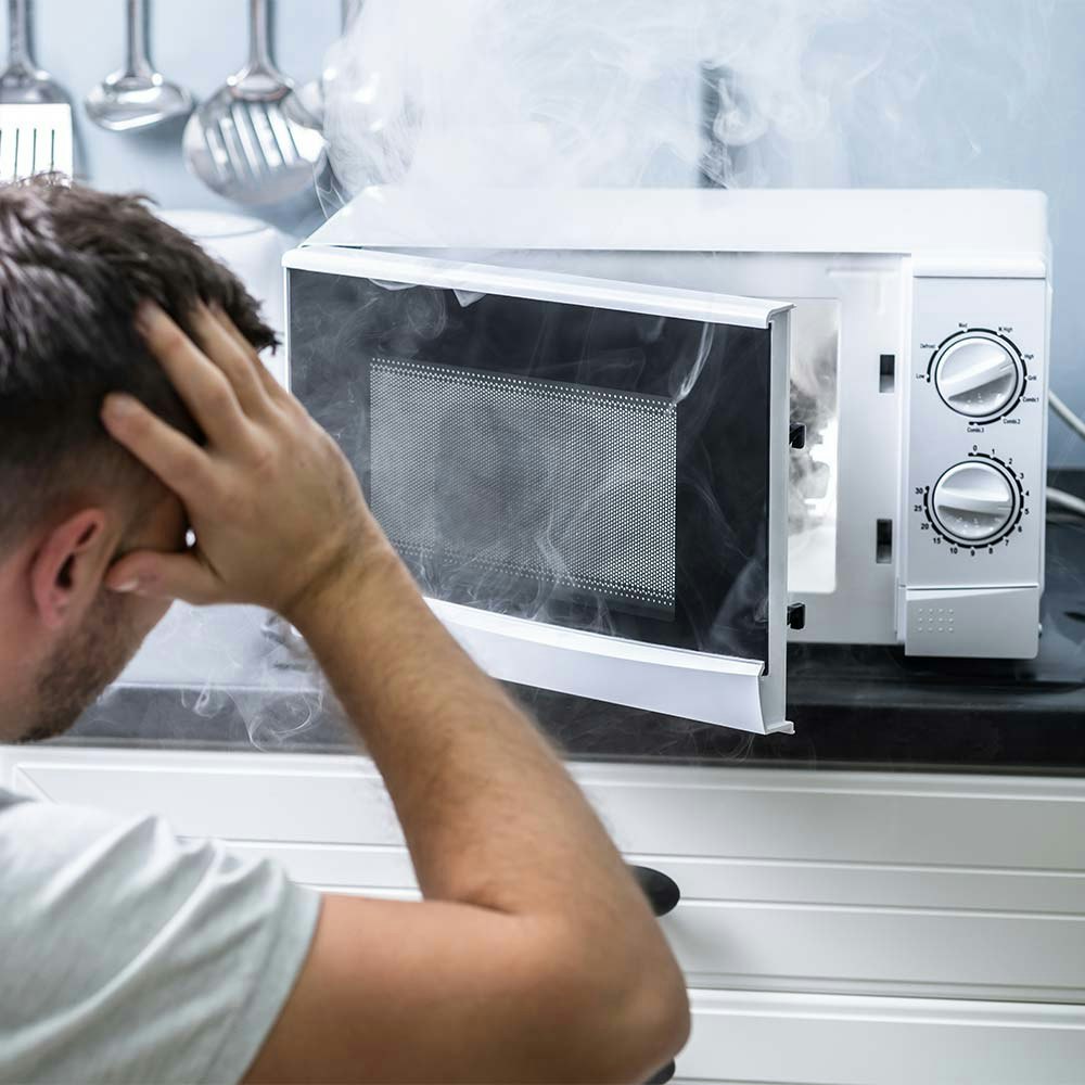 Man clutching head looking at microwave with smoke