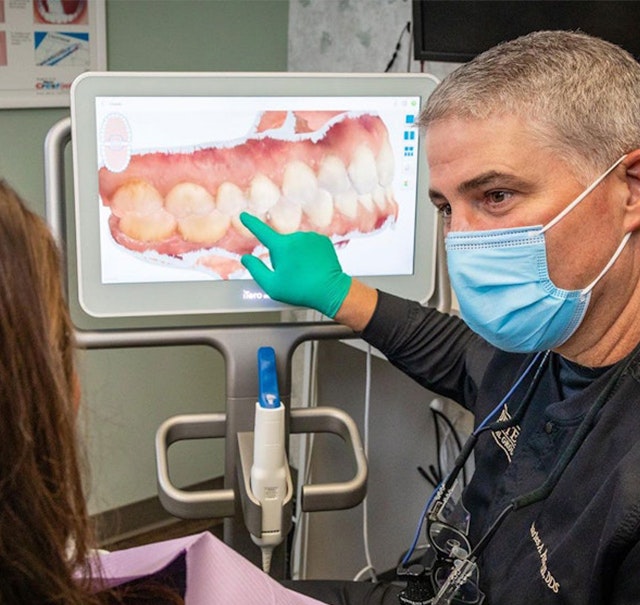 Dr. Charles Porter wearing a face mask and gloves, pointing to a digital image of a patient’s teeth on a monitor during a consultation in a dental office.