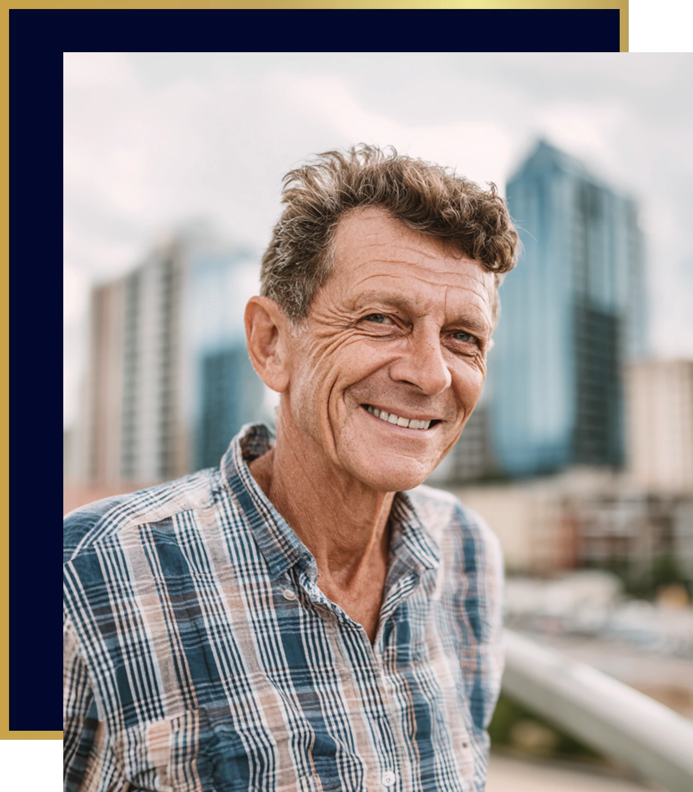 Smiling older man wearing a plaid button-down shirt standing outdoors with a city skyline blurred in the background.