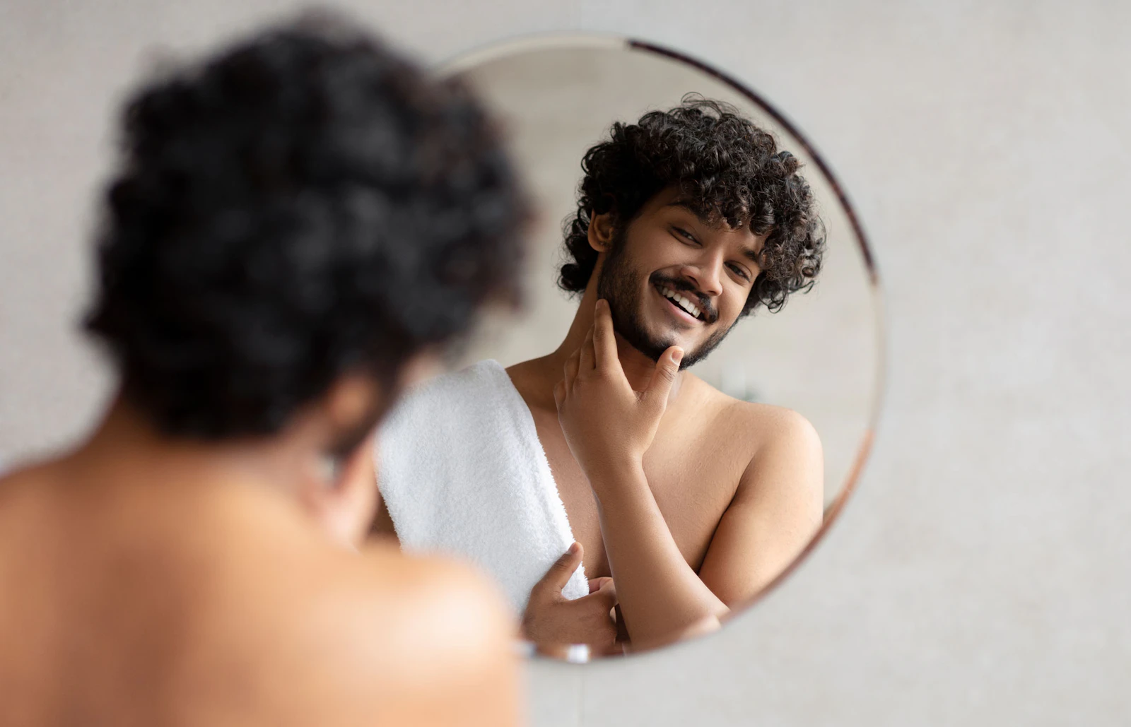 Man with curly hair smiling and checking his facial hair in a round mirror