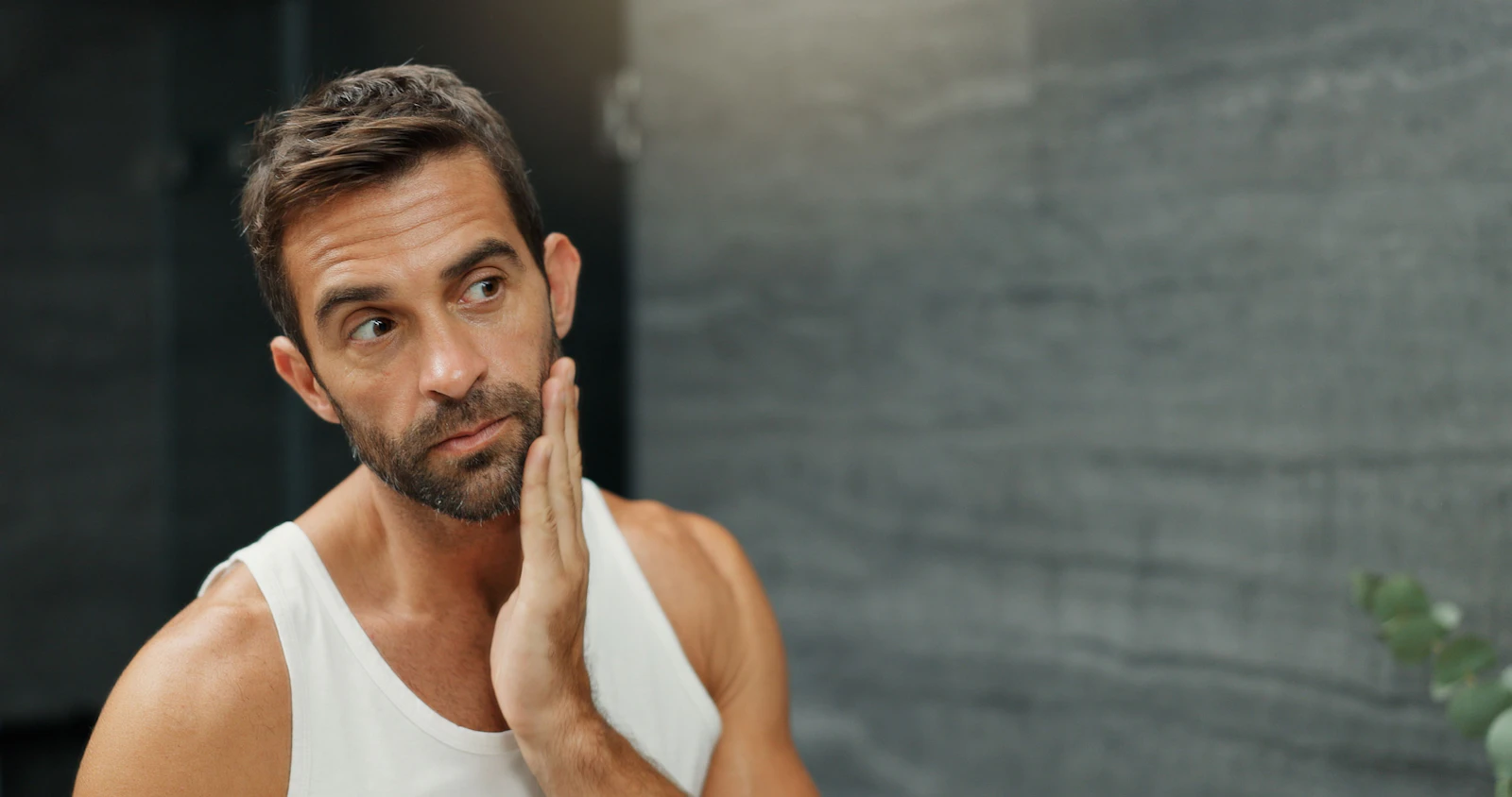 Man examining uneven stubble along his cheek and jaw in a bathroom mirror