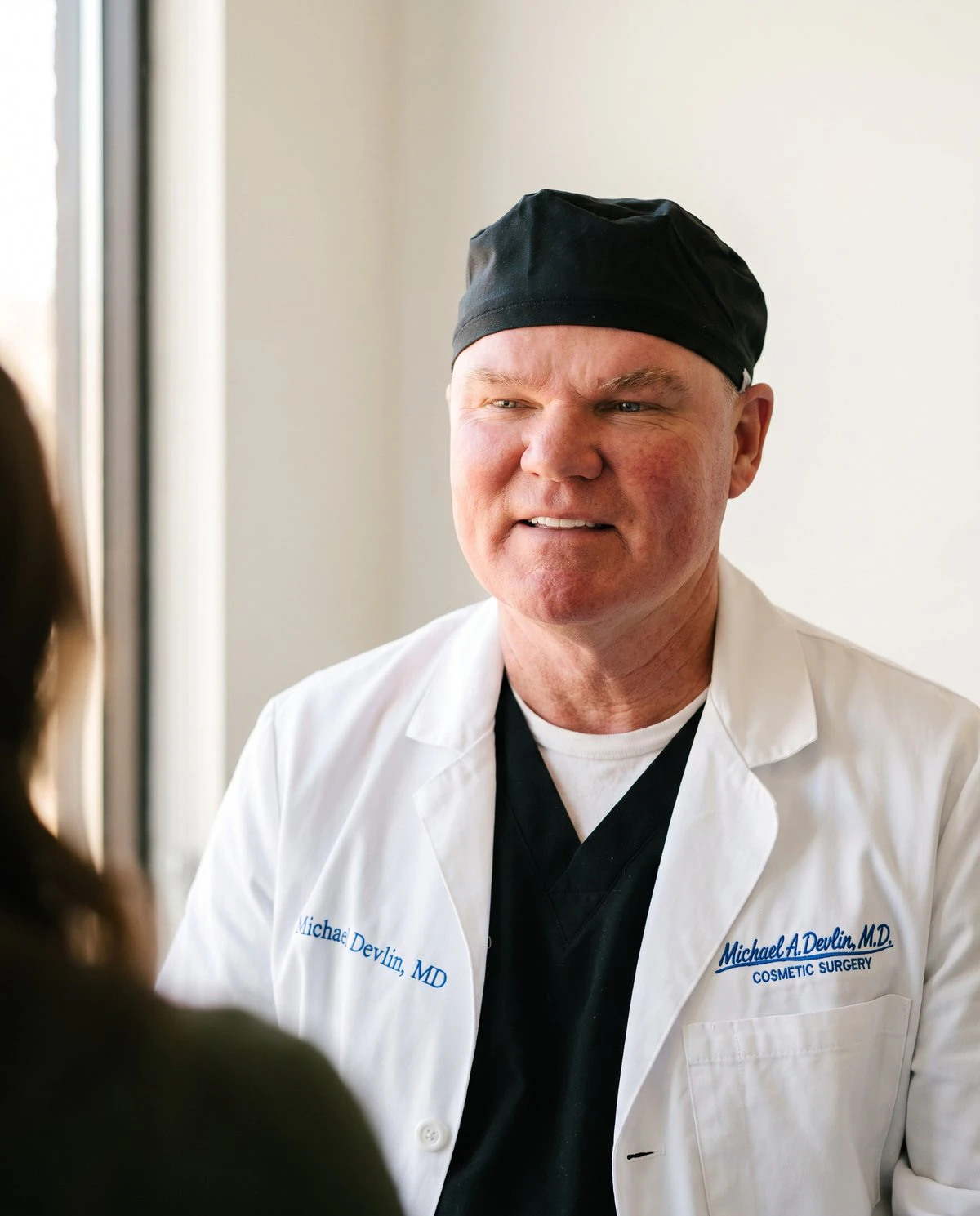 Dr. Michael Devlin in his white coat and black surgical cap, listening to a patient during a breast augmentation consultation