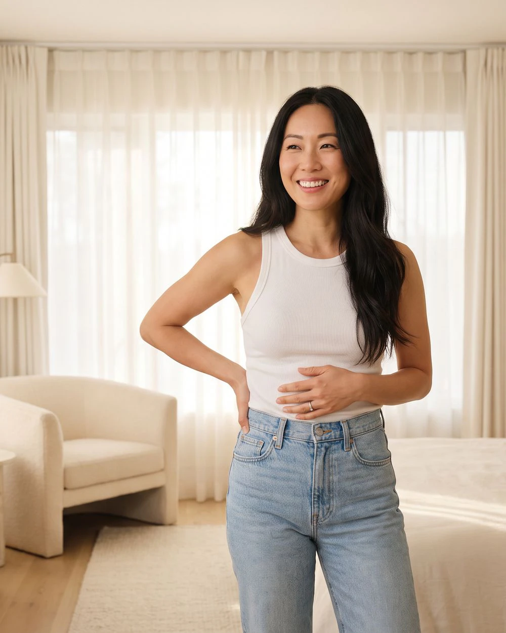 A smiling woman in a white tank top and jeans stands in a bright bedroom with one hand on her stomach