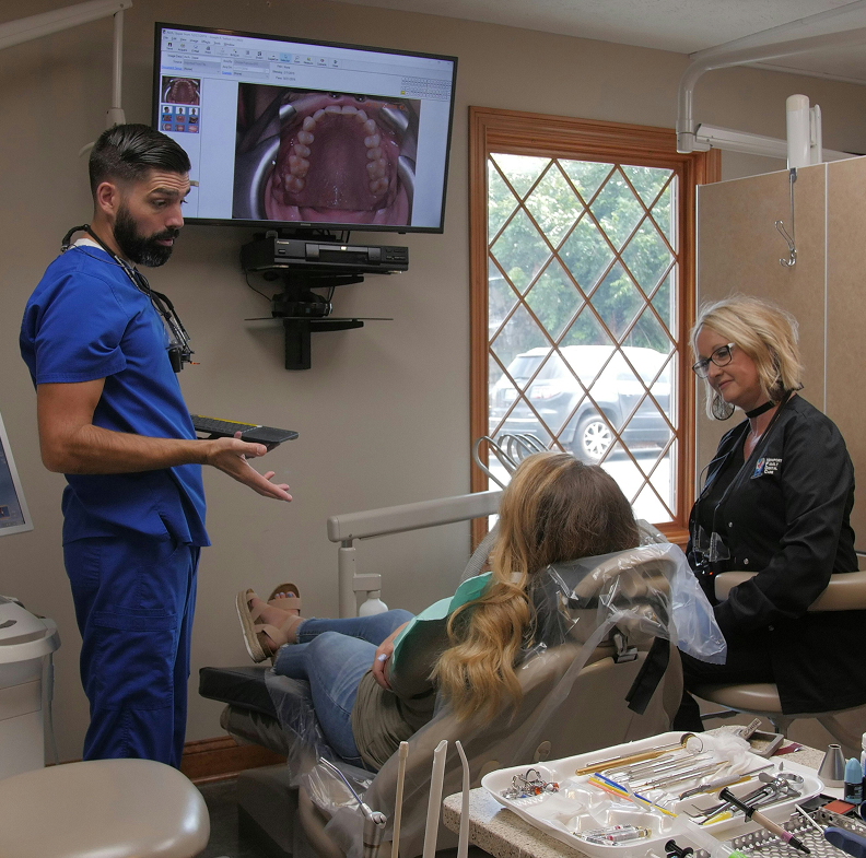 Dr. Joseph Sutton in blue scrubs reviewing dental images and speaking with a patient during an exam, with a team member seated nearby