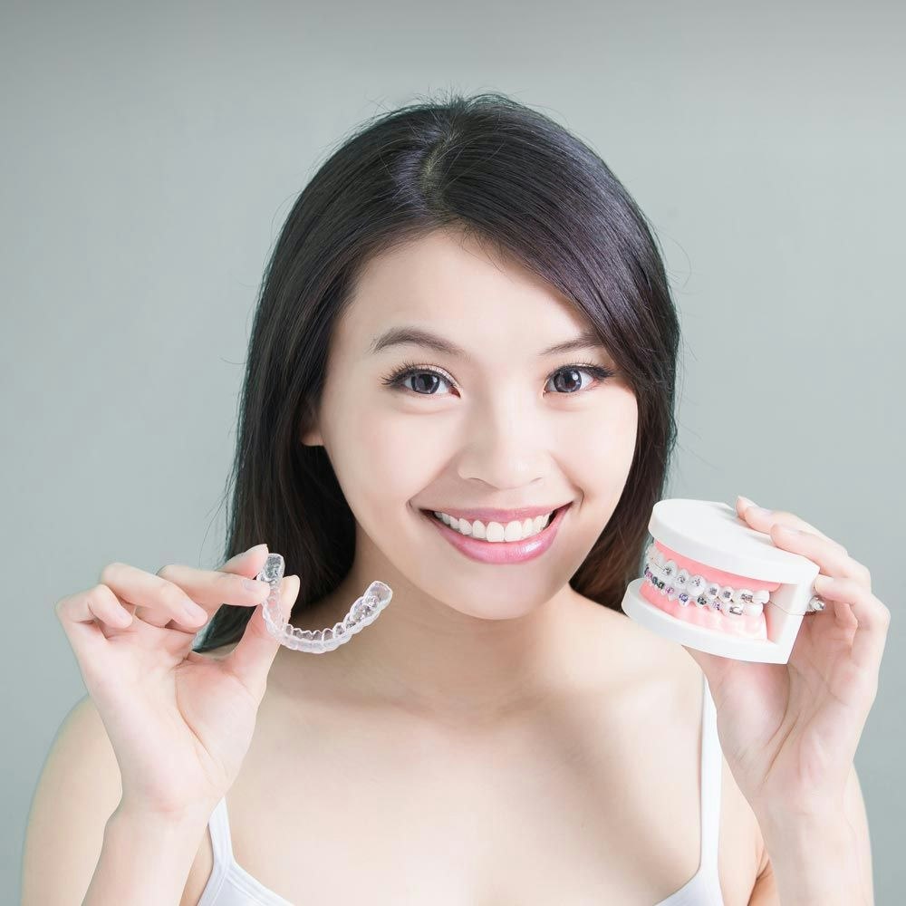 Smiling woman holding up aligner tray and model of teeth with metal braces