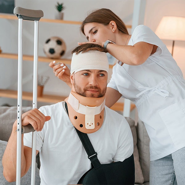 Man in neck and arm brace at home with a nurse wrapping bandages around his forehead