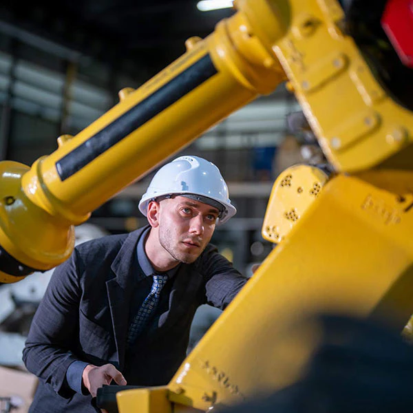 Man in a hard hat and suit leaning against a yellow industrial robotic arm