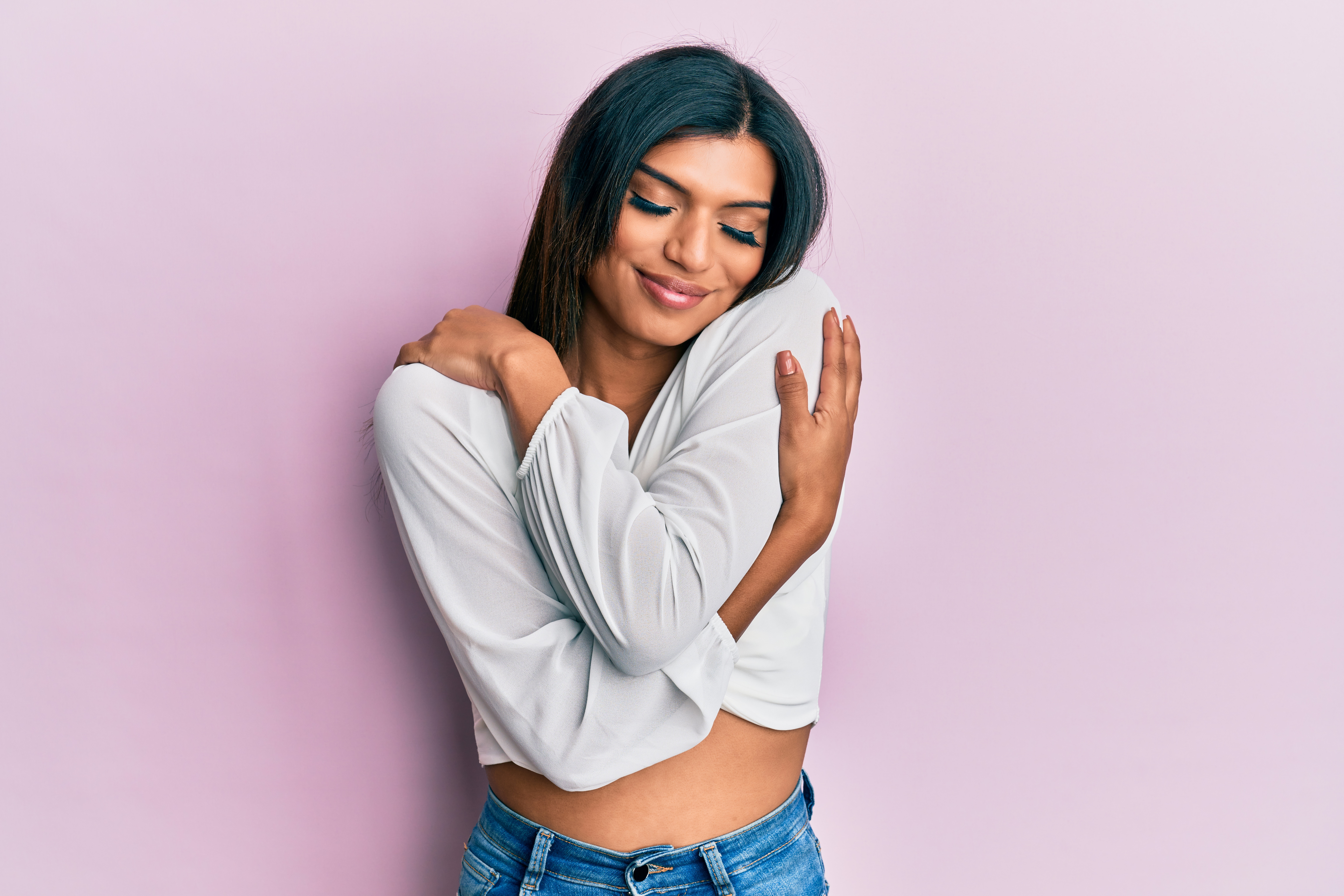 Smiling woman in a white top and jeans hugging herself against a pink background