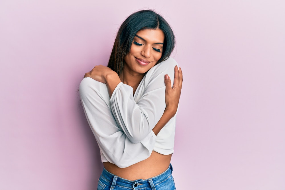 Smiling woman in a white top and jeans hugging herself against a pink background