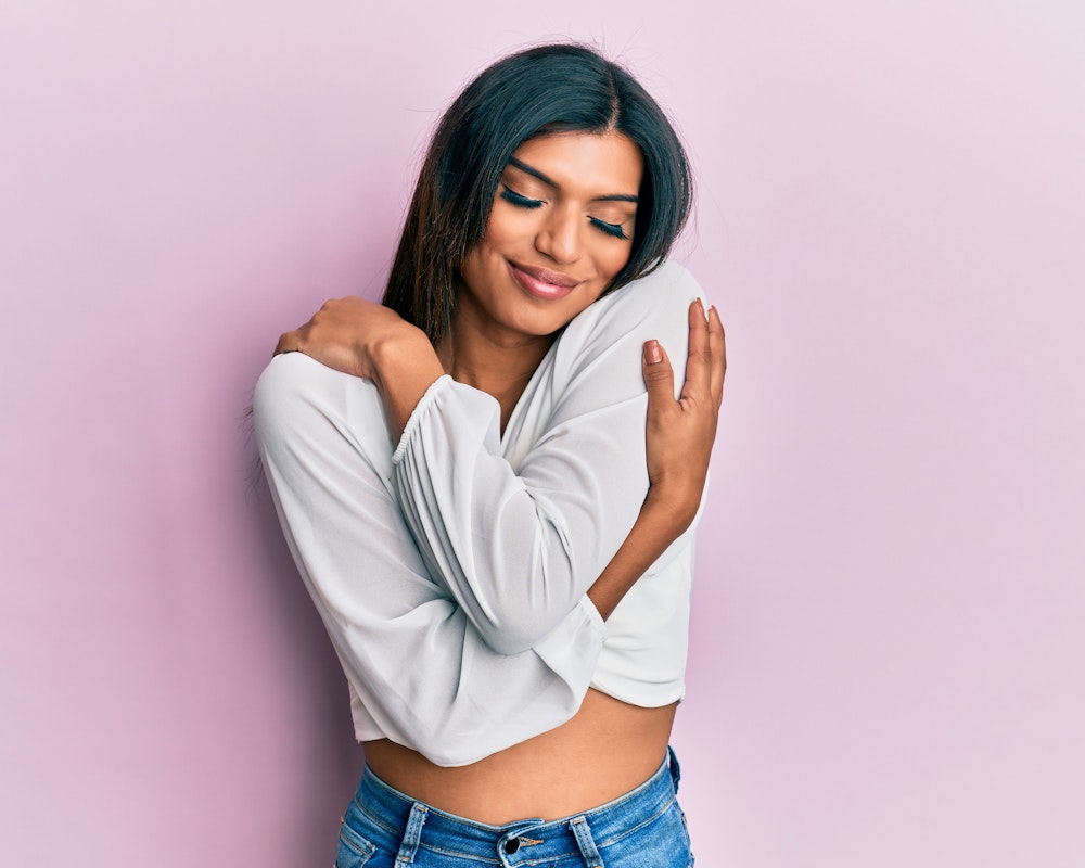 Smiling woman in a white top and jeans hugging herself against a pink background