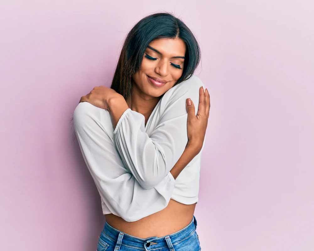 Smiling woman in a white top and jeans hugging herself against a pink background