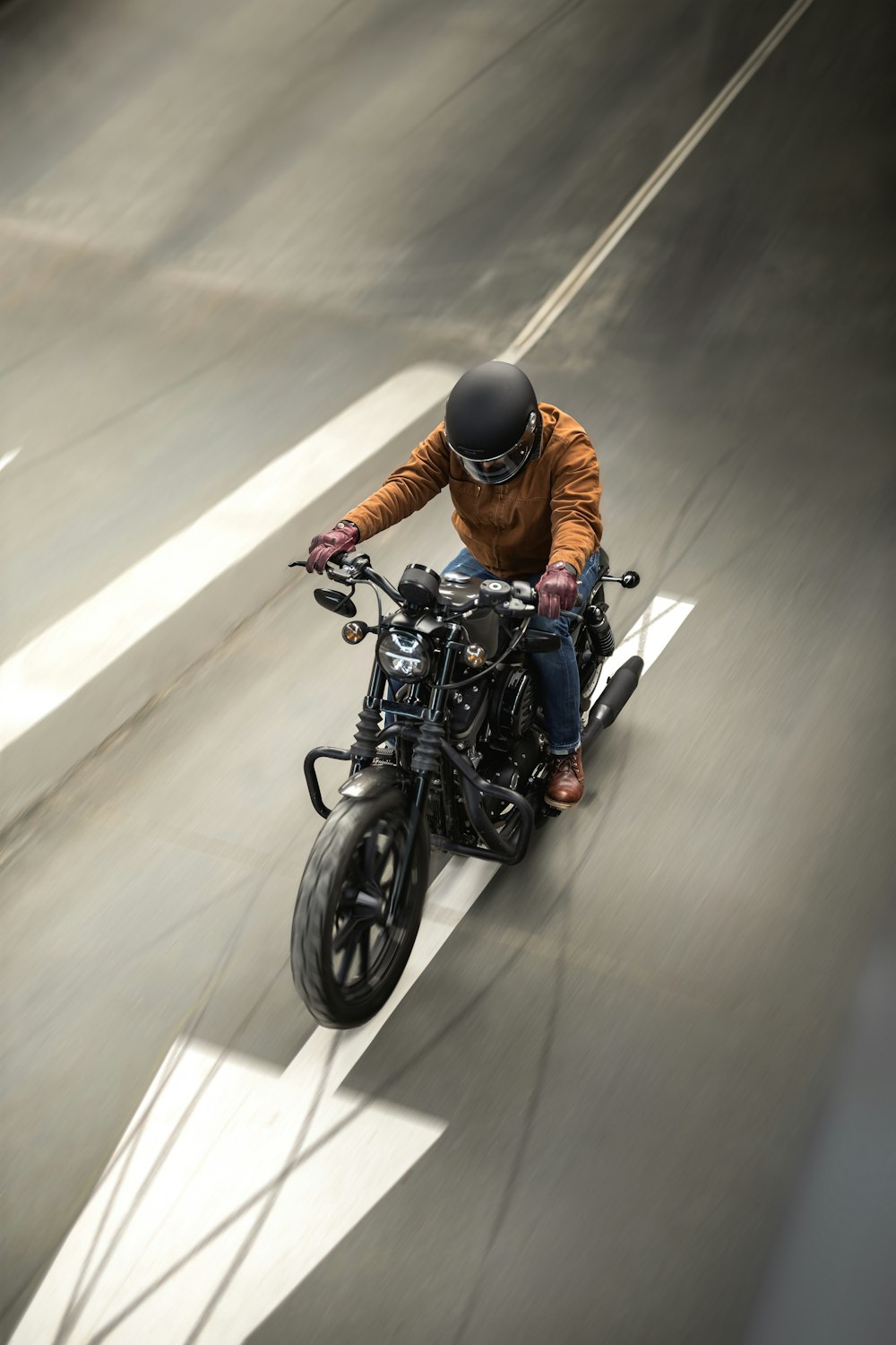 Motorcyclist wearing a helmet and brown jacket riding a black motorcycle down a city street.
