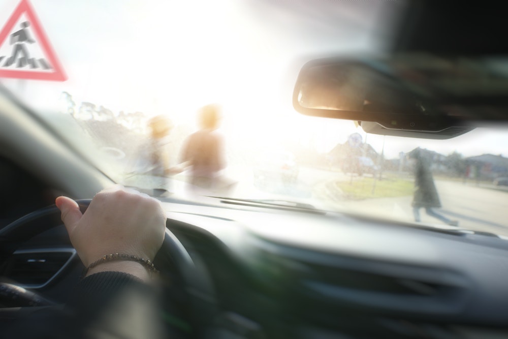 Driver’s perspective through a windshield at dusk: pedestrian blur in crosswalk ahead, car dashboard visible.