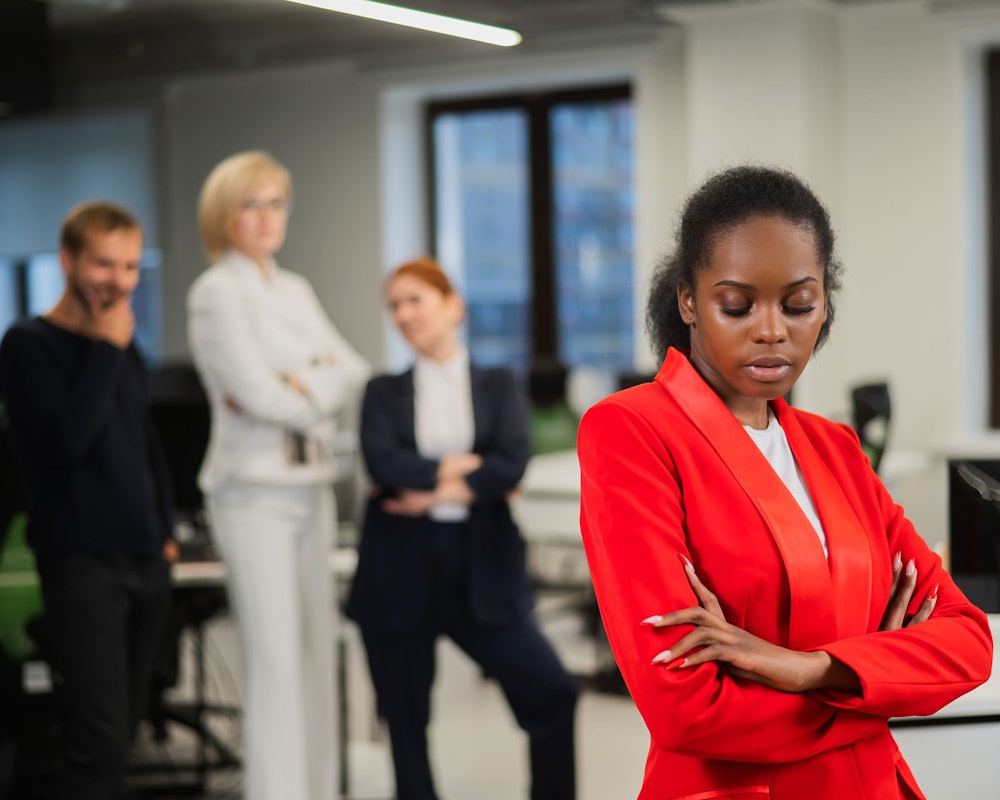 Black woman with her arms crossed while two coworkers talk in the background