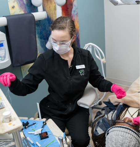 Female dental hygienist wearing pink gloves preparing tools while treating a patient covered in a blanket, inside a colorful exam room
