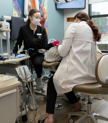 Two female dental professionals working on a patient in a modern exam room, with dental tools and equipment visible in the foreground