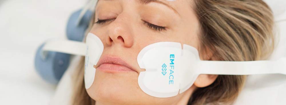 Woman with eyes closed receiving EMFACE treatment with applicator pads positioned on her cheeks