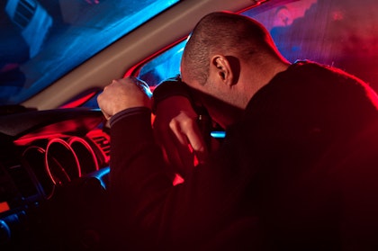 Man resting head on steering wheel, police lights in the background