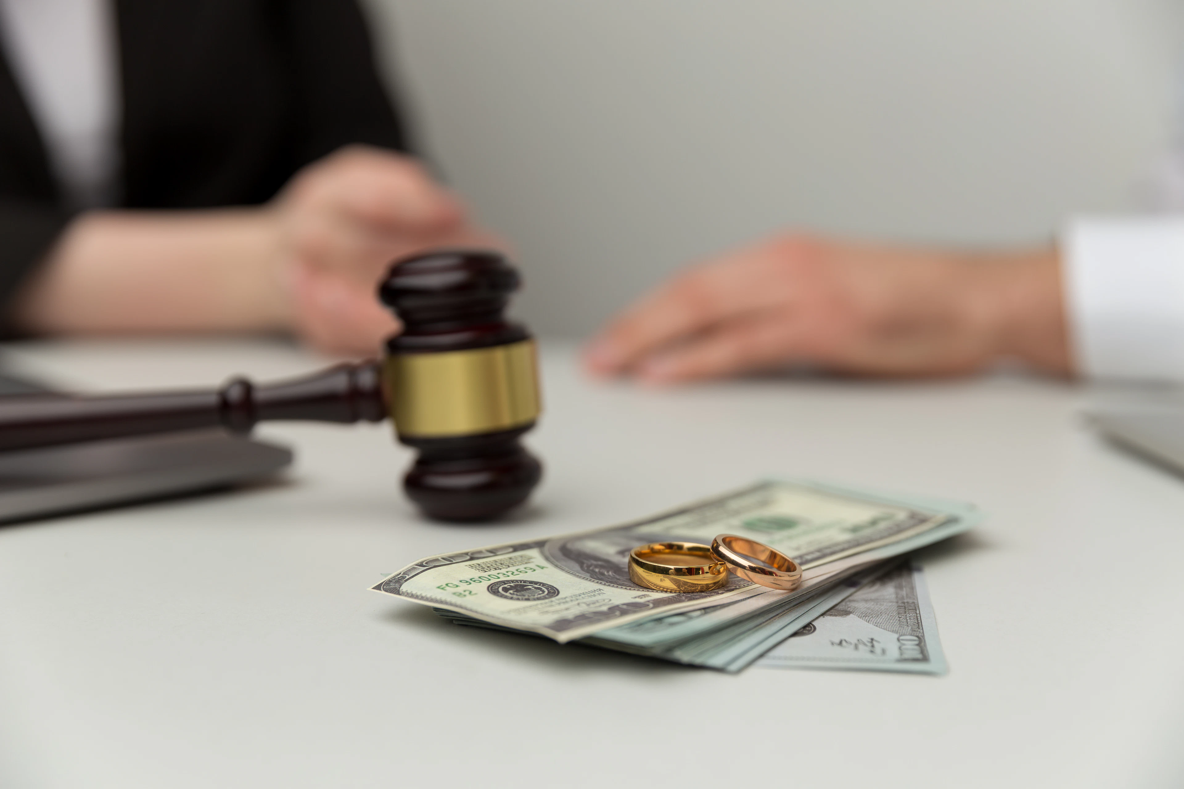 Money and wedding rings on a desk with gavel and people having a conversation in the background