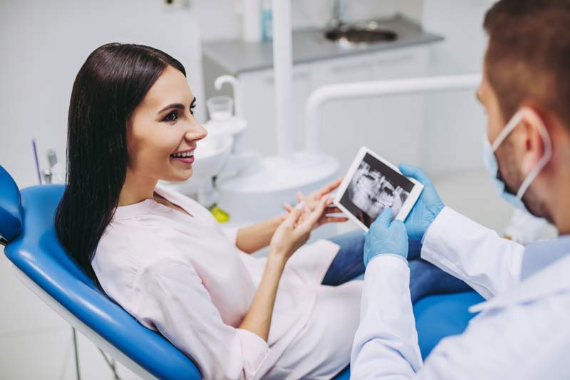 Dentist showing dental X-ray on tablet to female patient seated in exam chair