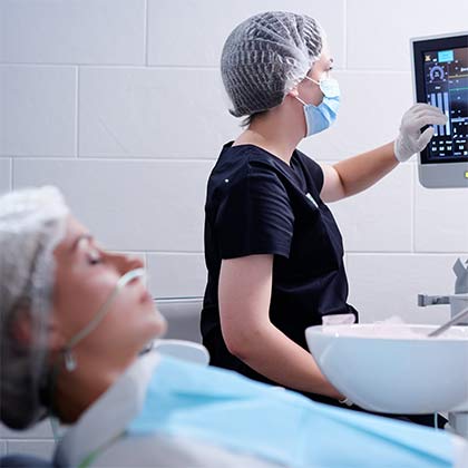 Dental professional in scrubs and mask monitoring a patient's vitals on a screen during a sedation dentistry procedure