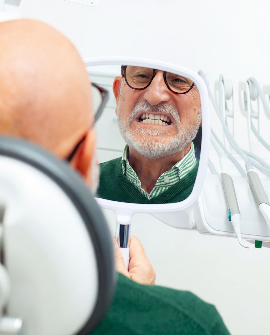 older man looking at teeth in hand mirror