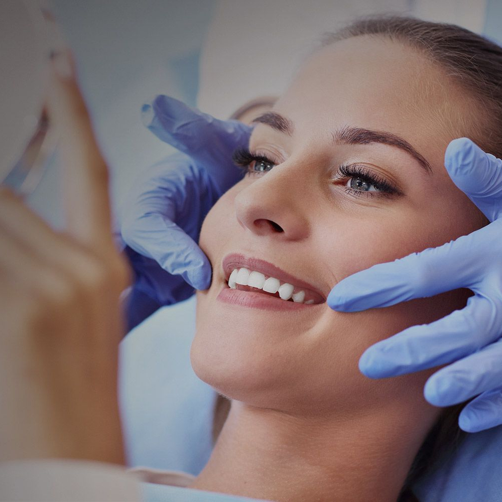 Woman looking at her smile in the mirror while the dentist's gloved hands hold her head