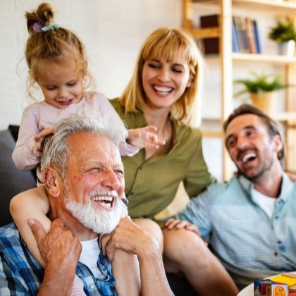Small child sits on grandfather's shoulders while mother and father laugh in background