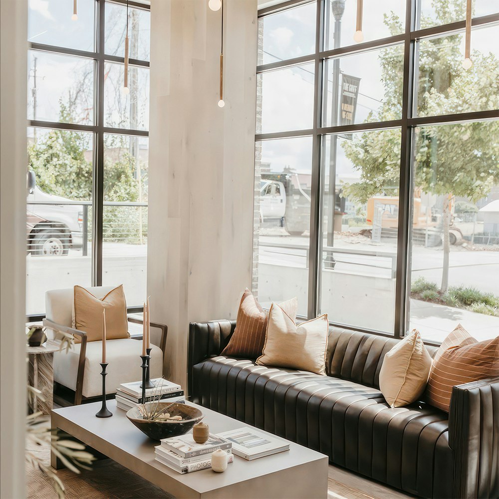Modern dental office lobby featuring a black leather sofa, tan accent pillows, white armchair, and coffee table with candles and books beside tall floor-to-ceiling windows.
