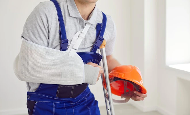 Construction worker in overalls with an arm in a sling holding a hard hat