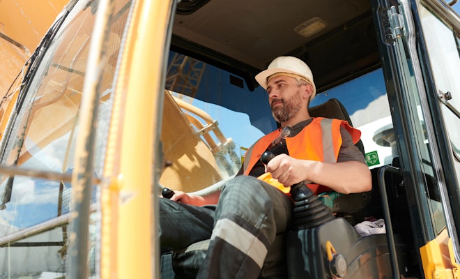 Construction equipment operator in a hard hat and safety vest seated inside a heavy machinery cab