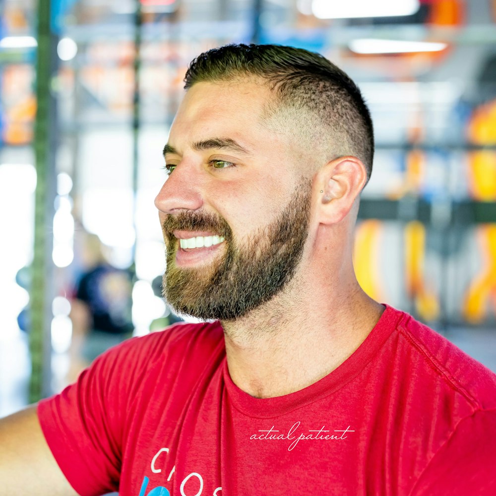 Smiling bearded man in a red shirt looking to the side in a bright indoor setting