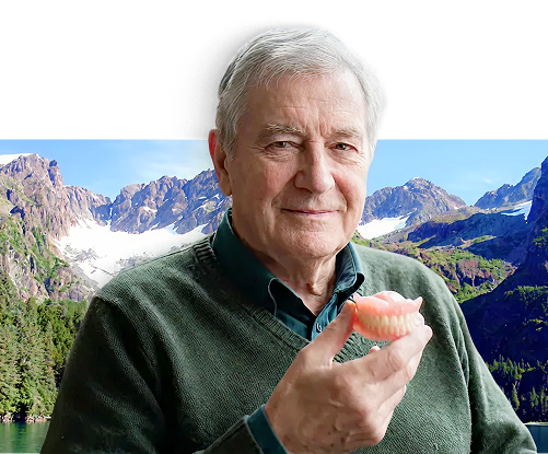 senior man holding up dentures with Alaska in the background