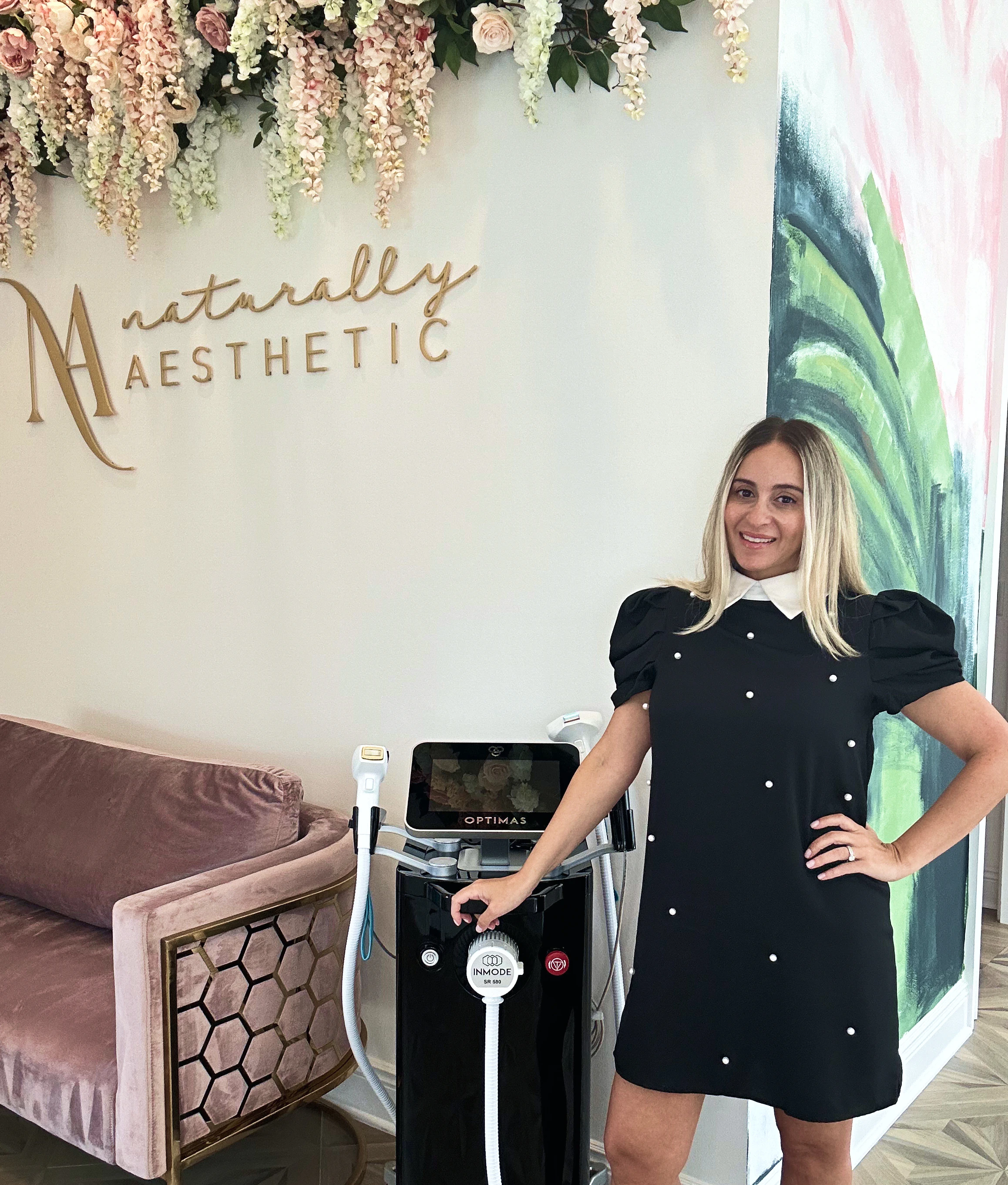 Nichole Gilbarty posing in front of Naturally Aesthetic wall and with OPTIMAS INMODE technology