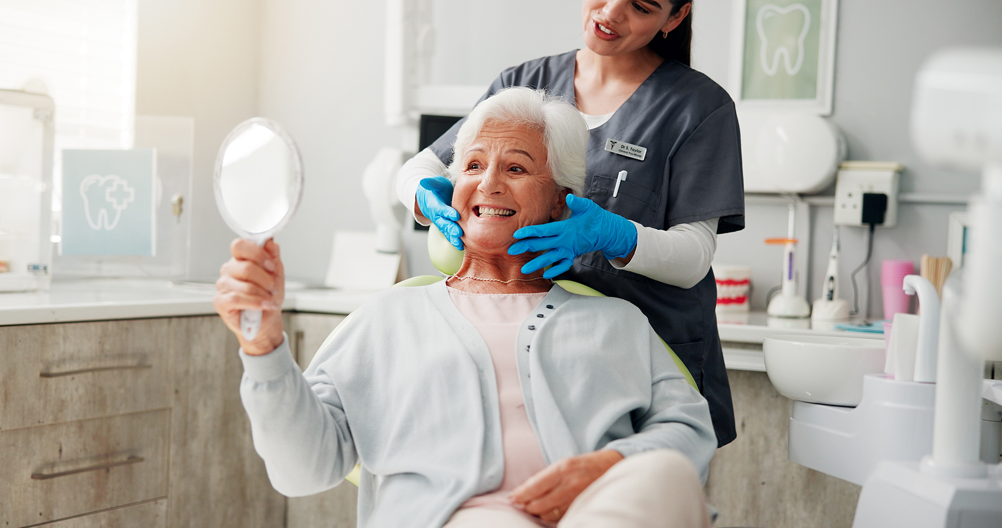 Older woman smiling while holding a mirror as a dental professional supports her jaw during a consultation.