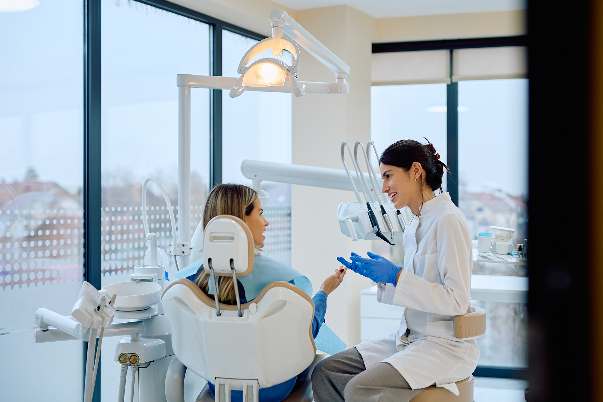 Dentist wearing blue gloves speaking with a patient seated in a dental chair in a bright exam room.