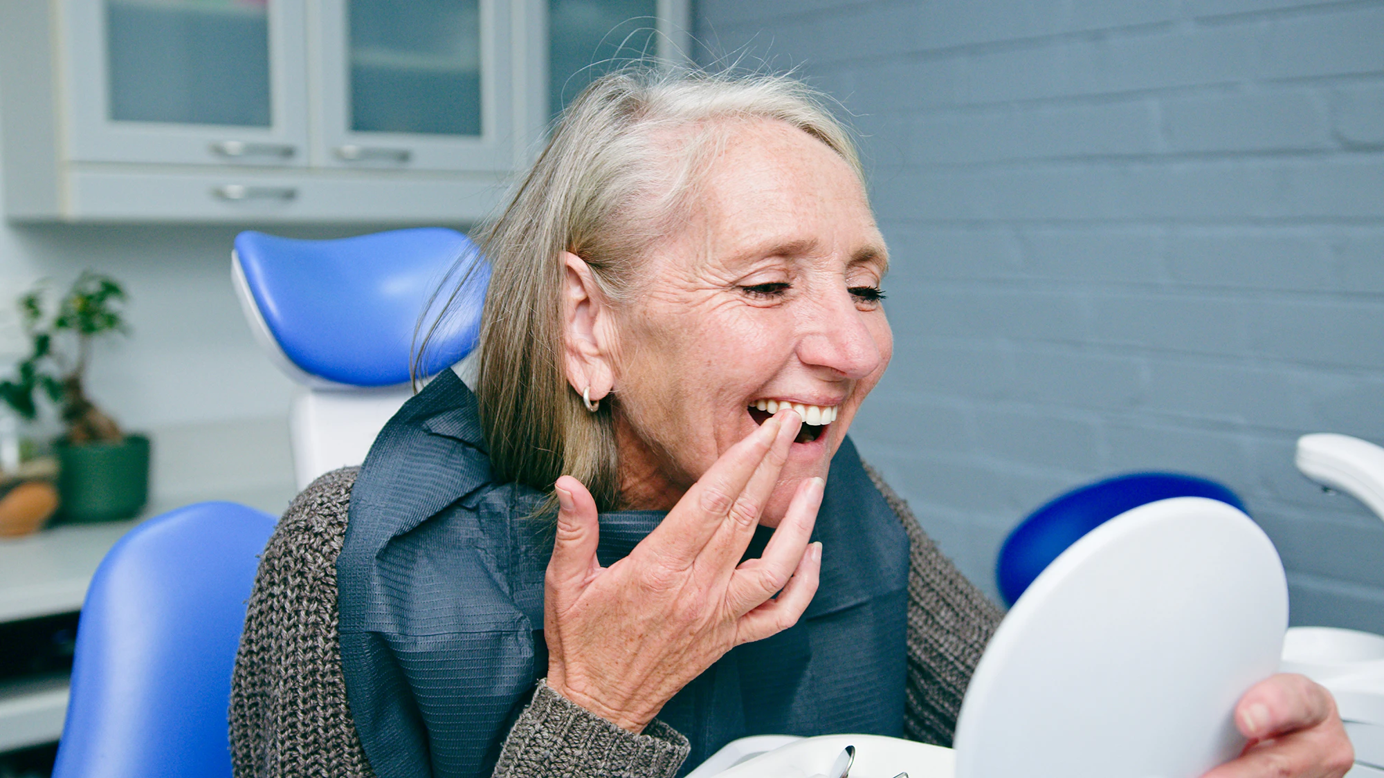 Older woman smiling while looking at her teeth in a handheld mirror while seated in a dental chair.