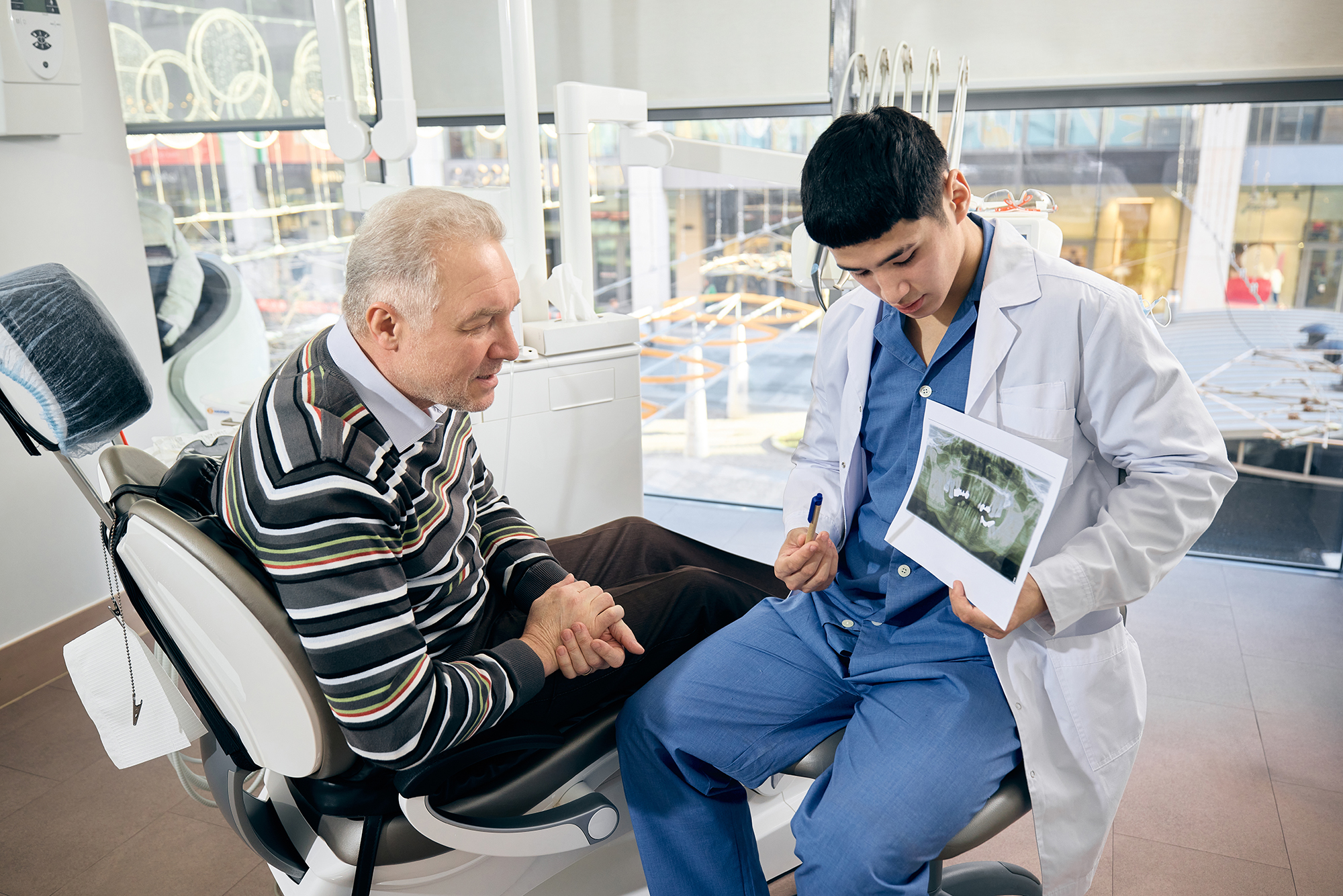 Dentist showing a dental image on a tablet while consulting with an older male patient in an exam room.