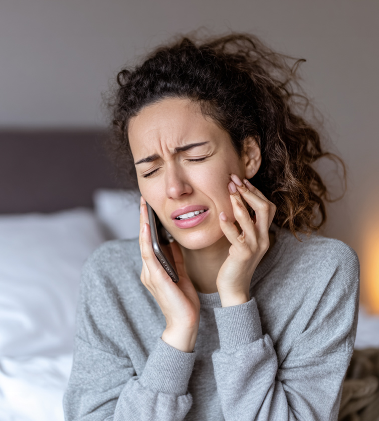 Woman holding her cheek in pain while talking on the phone, seated on a bed.