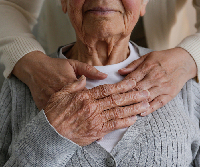 Elderly woman sitting with younger caregiver’s hands resting gently over hers on her chest, conveying support and connection