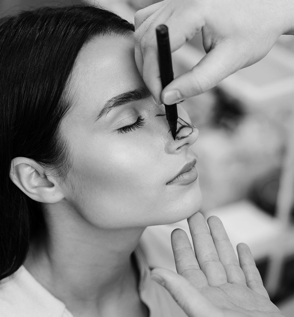Close-up of a woman with eyes closed while a clinician marks surgical guidelines on her nose with a pen during a rhinoplasty consultation.