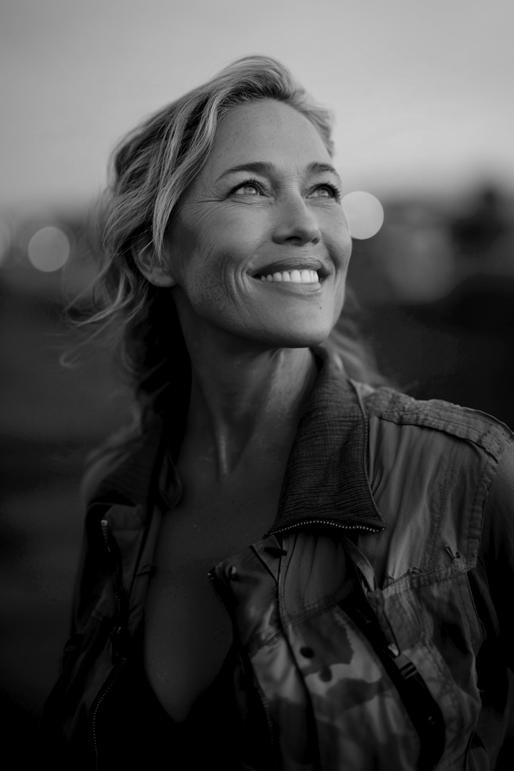 Smiling middle-aged woman outdoors at dusk, looking upward with a bright, refreshed expression.