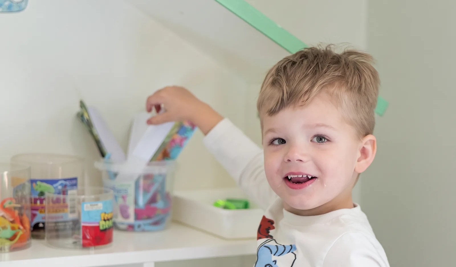 A young boy smiling over his shoulder while choosing a toy from the play shelf.